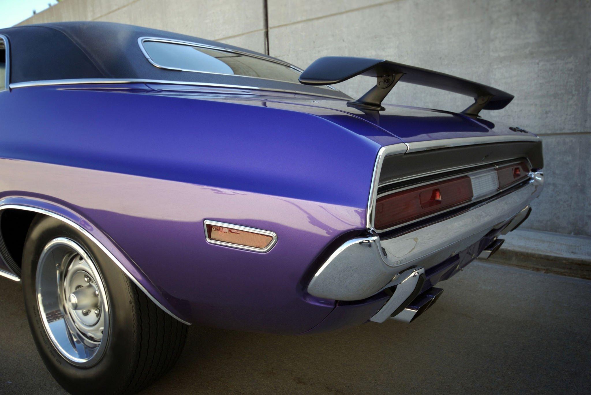 A shiny purple vintage car with a black vinyl roof, chrome accents, and a rear spoiler, parked against a textured concrete wall.