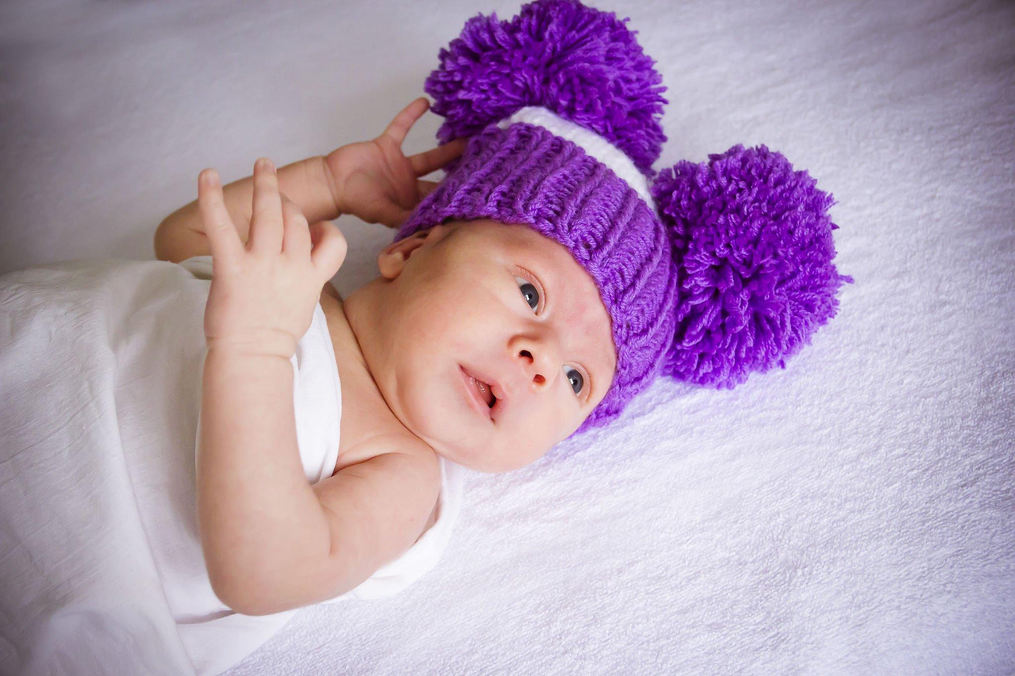 A baby lies on a white blanket, dressed in a white outfit and wearing a vibrant purple knitted hat with two fluffy pom-poms.